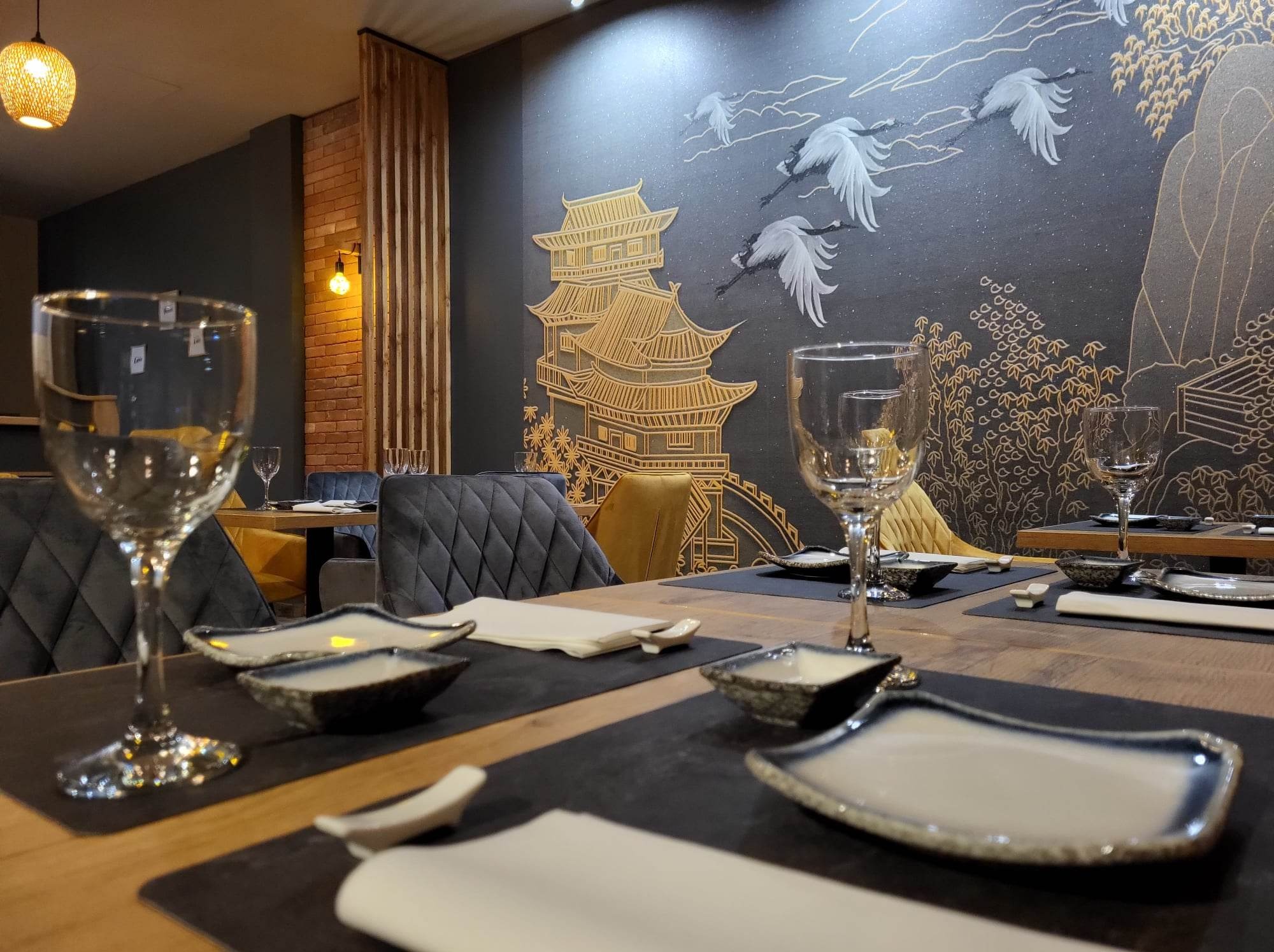 Elegancko nakryty stół w restauracji z motywami orientalnymi na ścianie. Widoczne talerze, szklanki i dekoracje w stylu japońskim.