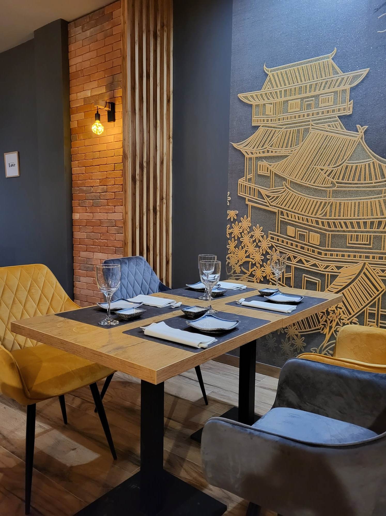 Ustawiony stół w restauracji z dekoracyjną ścianą w stylu azjatyckim. Żółte i szare krzesła, nakrycia i grafika budynku na ścianie tworzą spójny wystrój wnętrza.