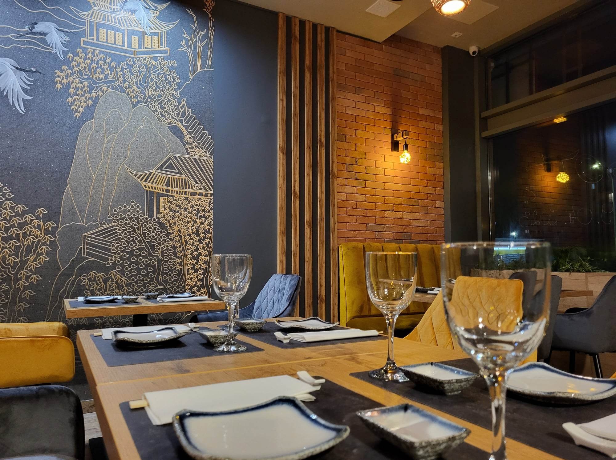 Stylowa aranżacja wnętrza restauracji z motywami azjatyckimi: dekoracyjna ściana z rysunkami, ceglana ściana, drewniane lamele i nakryty stół z zastawą.