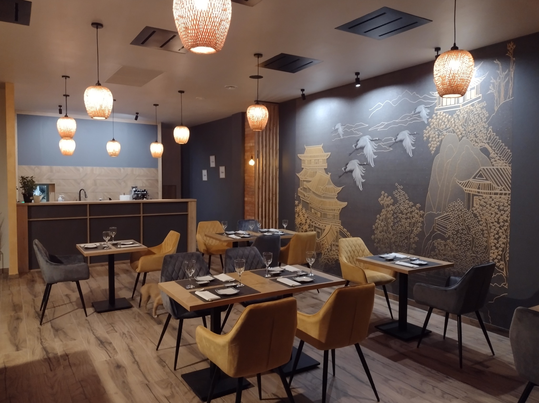 Eleganckie wnętrze restauracji w stylu orientalnym. Ciemne ściany zdobi złoty rysunek pagody i żurawi. Drewniane stoły nakryte dla gości, rattanowe lampy.