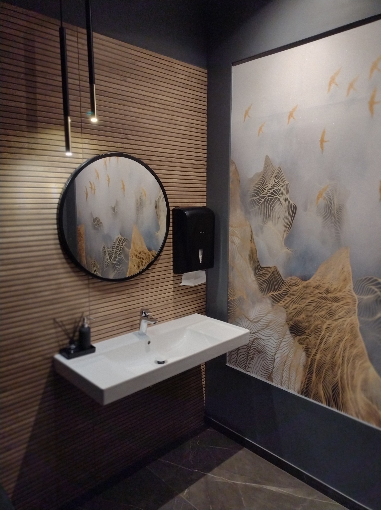 Nowoczesna łazienka z umywalką, okrągłym lustrem i dekoracyjną ścianą z motywem gór i ptaków. Minimalistyczne oświetlenie i ciemna kolorystyka dodają elegancji.