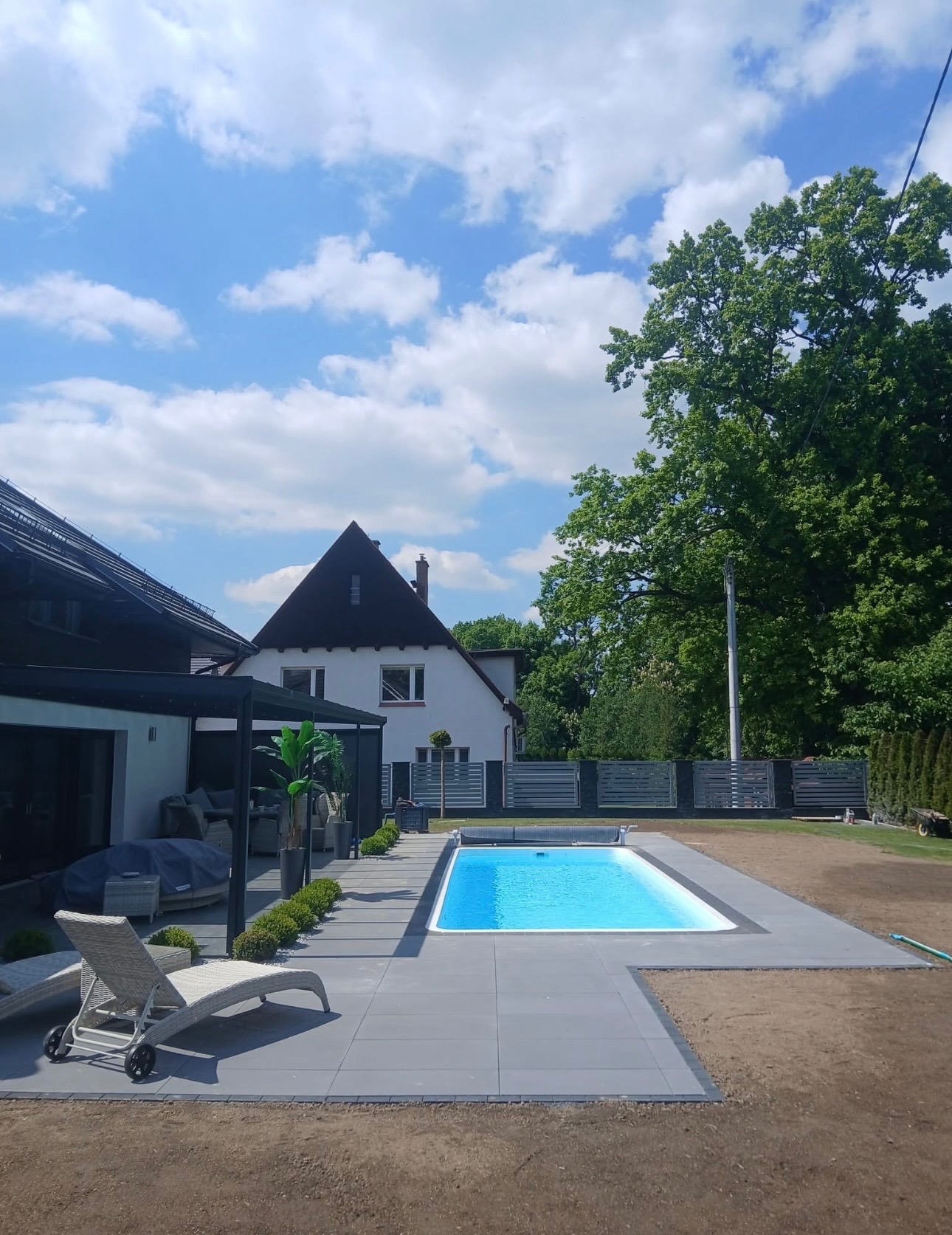 Nowoczesny basen otoczony szarą kostką brukową, z leżakiem i pergolą w tle, na tle domu i zieleni. Słoneczny dzień, błękitne niebo z chmurami.
