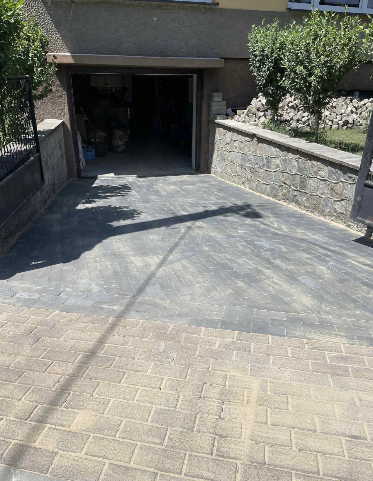 Ułożona szara kostka brukowa prowadząca do garażu z kamiennym murem oporowym i stertą kamieni w tle. Część nawierzchni wykończona jaśniejszą kostką.