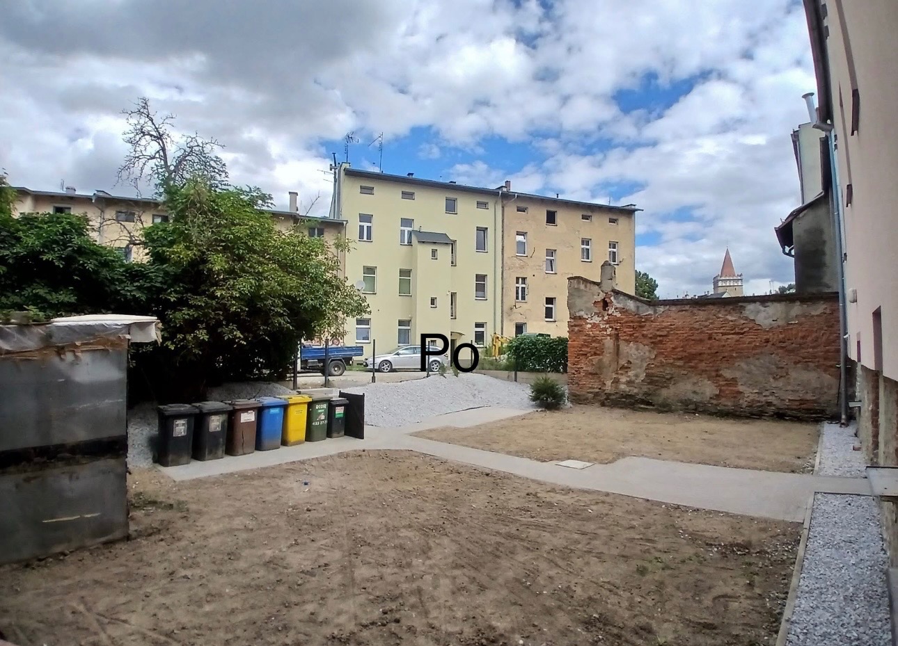 Ukończona ścieżka z kostki brukowej na podwórku kamienicy, widok z perspektywy ziemi, z boku widoczne pojemniki na śmieci i gruz.