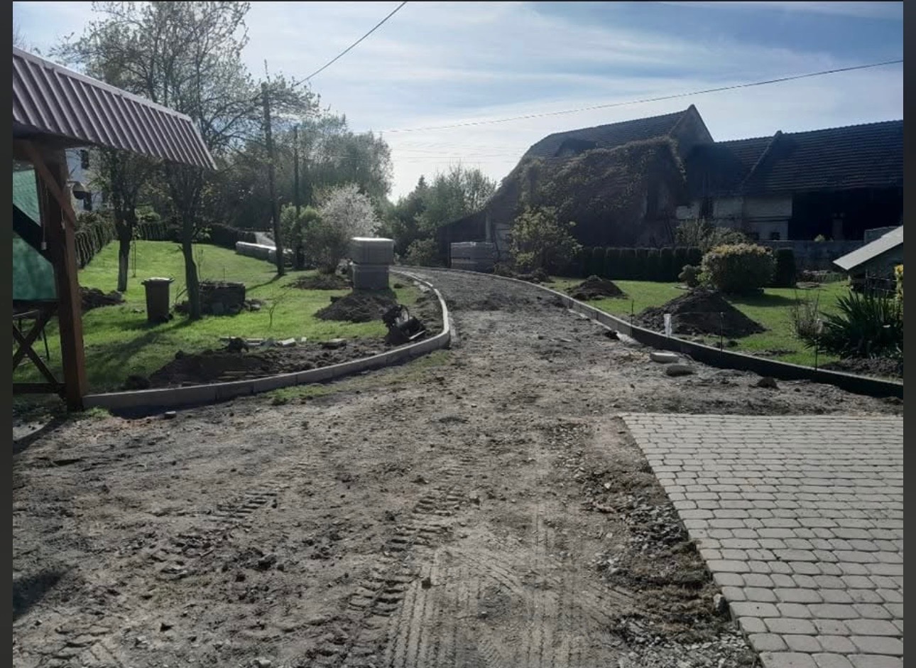 Kształtowanie terenu podjazd z obrzeżami, widoczne ślady opon, w tle budynki gospodarcze i zieleń. Częściowo ułożona kostka brukowa po prawej stronie.