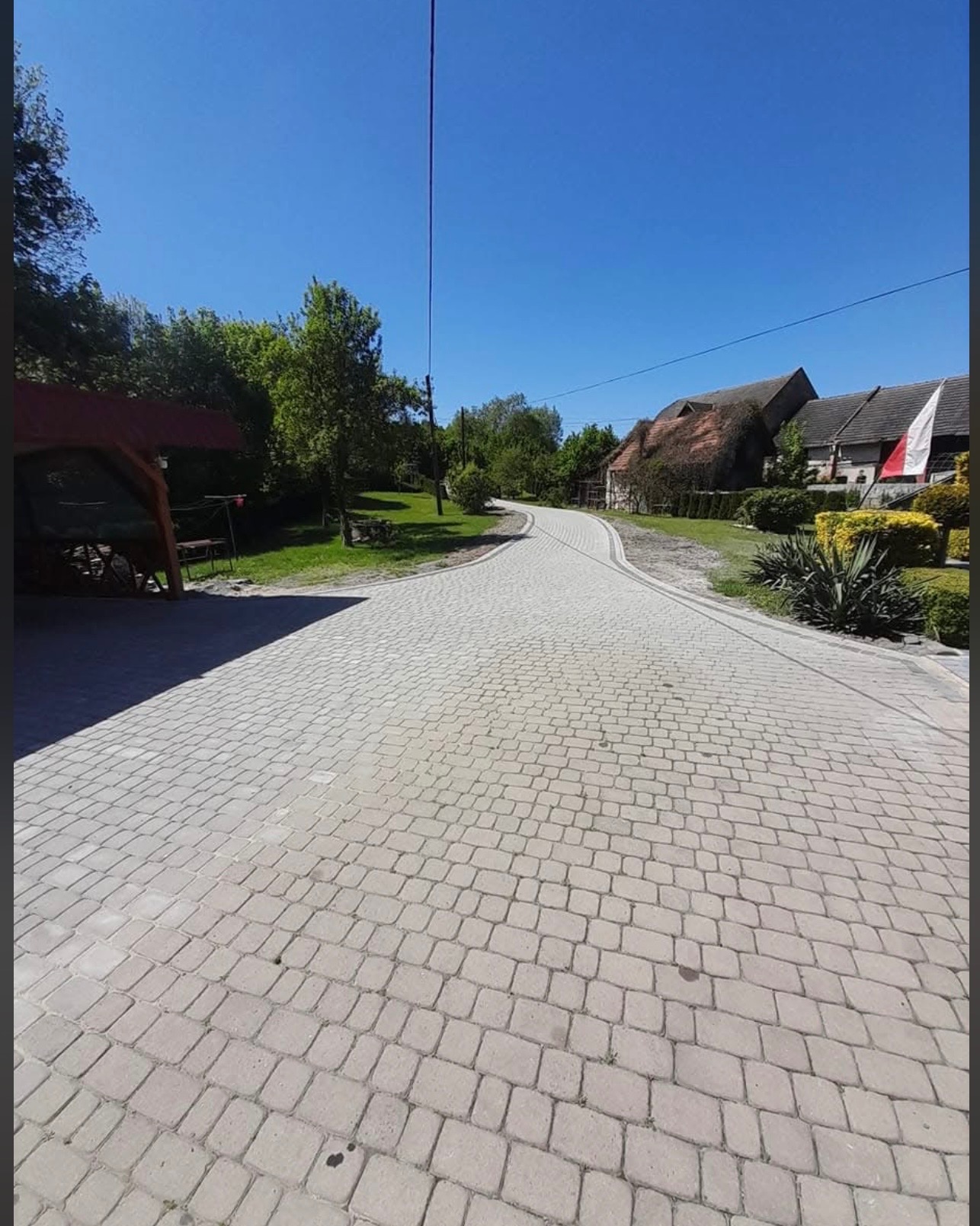 Długa, wijąca się ścieżka z jasnoszarej kostki brukowej prowadzi w kierunku budynków z dachami krytymi strzechą, otoczona zielenią i błękitnym niebem.