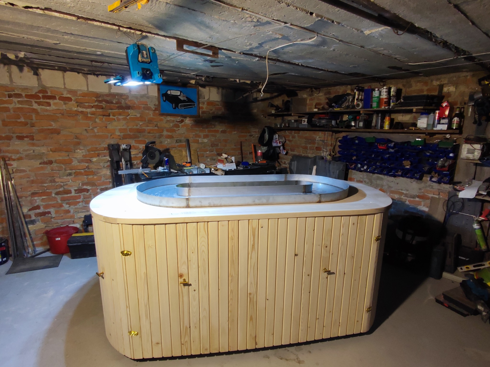 Drewniana obudowa wanny z metalowym wkładem, widoczna w warsztacie z ceglaną ścianą i narzędziami. Oświetlenie warsztatowe, surowy betonowy sufit.