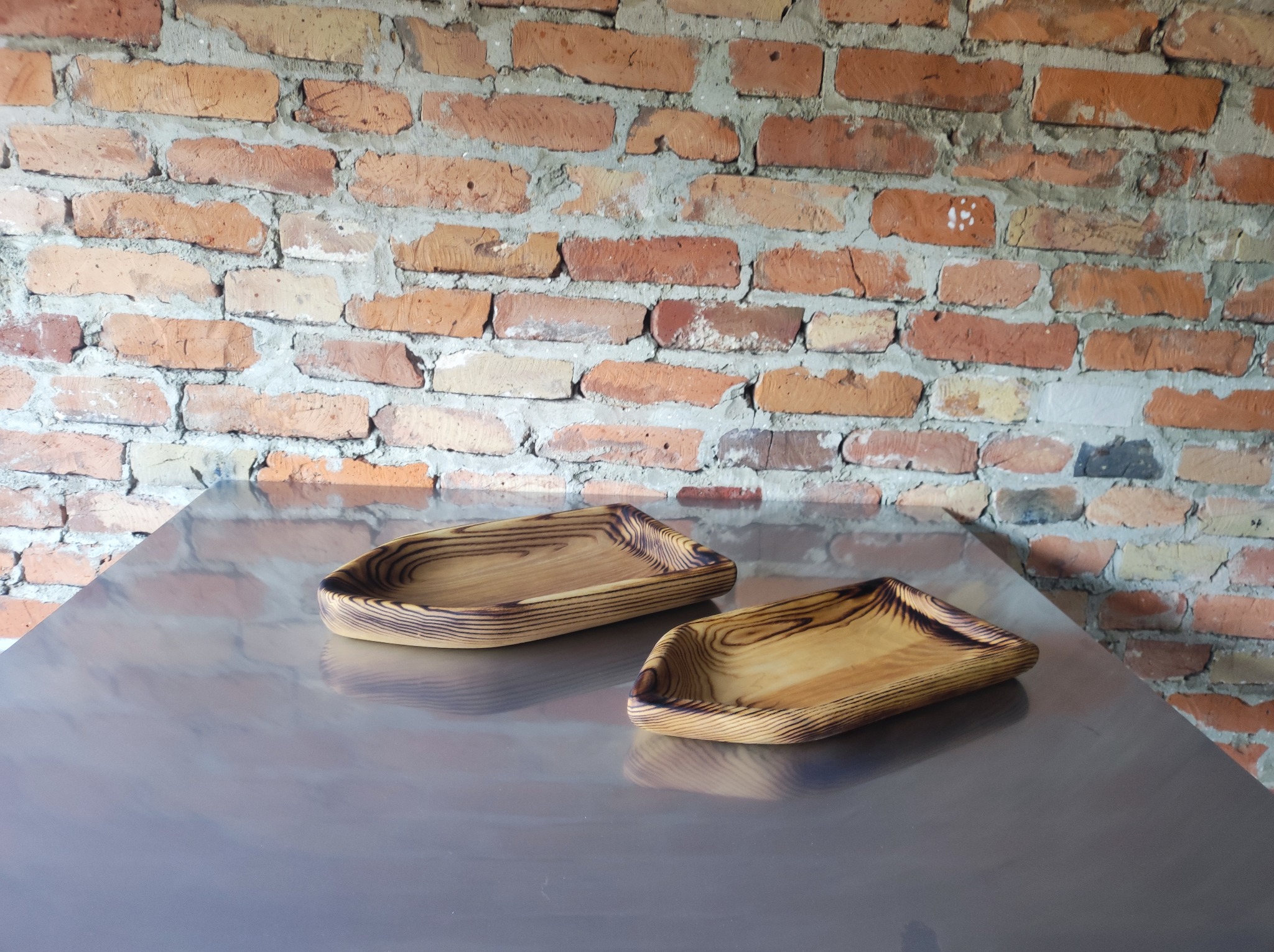 Dwie unikatowe, ręcznie robione drewniane miseczki z wyraźnym rysunkiem słojów, leżą na metalowym blacie na tle ściany z czerwonej cegły z widoczną zaprawą.