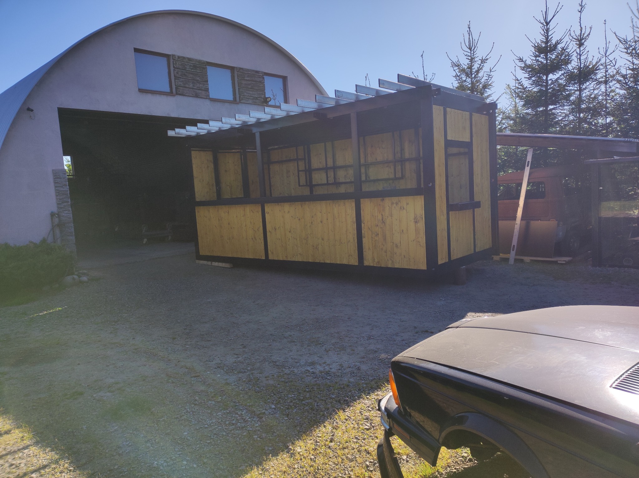 Drewniana pergola z metalowym stelażem na tle budynku gospodarczego i drzew iglastych w słoneczny dzień. Fragment samochodu na pierwszym planie.