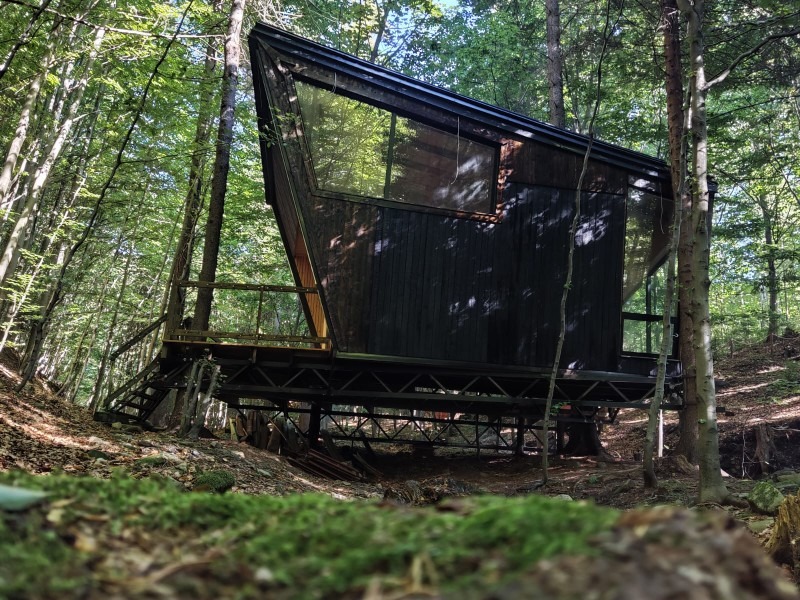 Nowoczesny, czarny domek letniskowy z dużymi oknami, wkomponowany w leśny krajobraz, wsparty na metalowej konstrukcji. Schody prowadzą na taras.