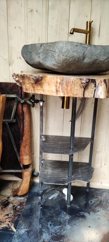 Unikatowa umywalka z kamienia na blacie z drewna z korą, wsparta na metalowym stelażu z półkami. Złota bateria, marmurowa podłoga, drewniane panele ścienne.