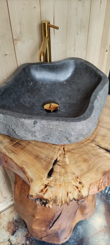 Kamienna umywalka o nieregularnym kształcie z złotą baterią na drewnianym blacie z widocznymi słojami, ustawiona przy ścianie z drewnianą okładziną.