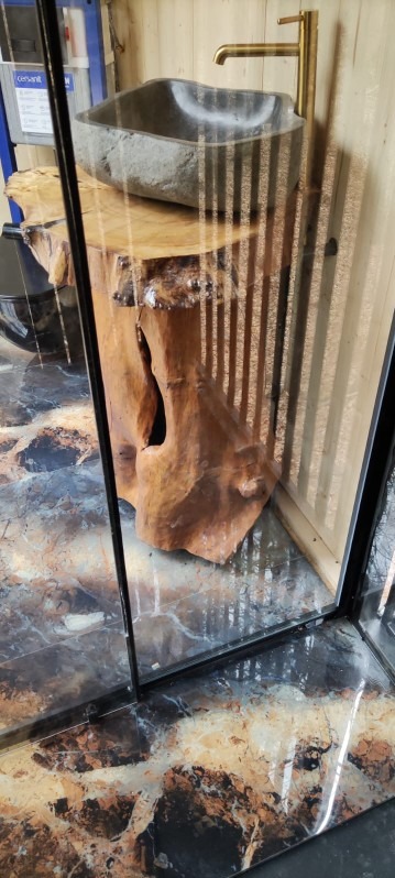 Łazienka z kamienną umywalką na drewnianym postumencie i złotą baterią, widoczna przez szklaną ścianę kabiny. Marmurowa podłoga.