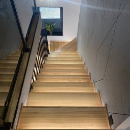 HolzForm Tomasz Jaśko - Nowoczesne schody drewniane z podświetleniem LED, czarne metalowe barierki, szara ściana z geometrycznym wzorem, widok z dołu, roślina w oknie na górze.
