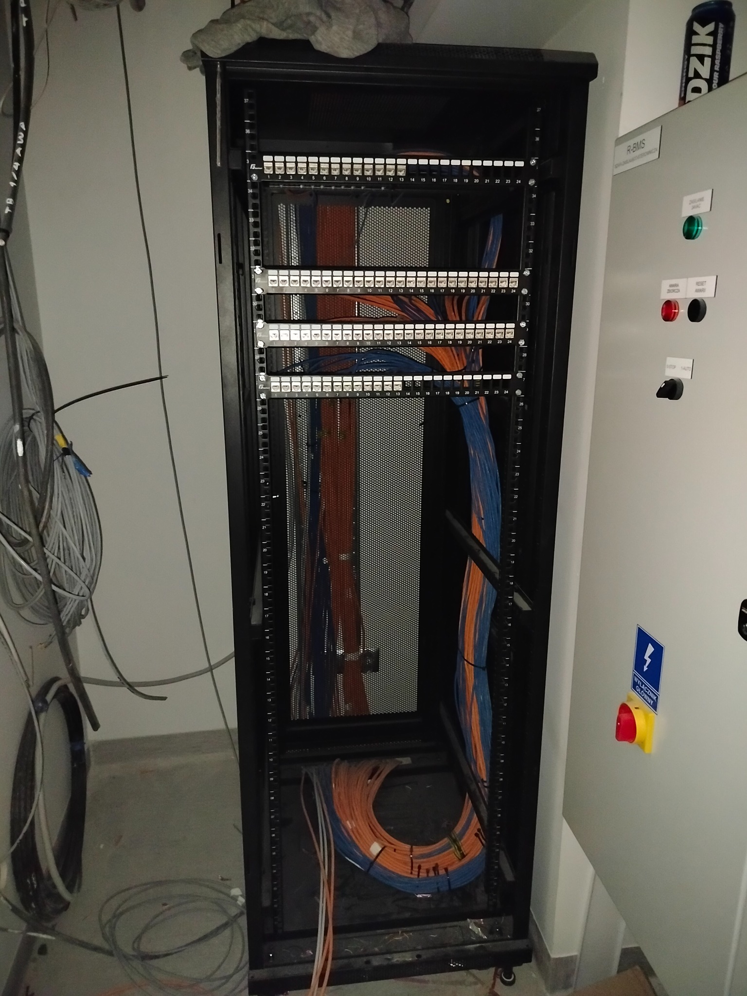 Szafa rack z panelami krosowniczymi i uporządkowanymi kablami Ethernet w kolorach pomarańczowym i niebieskim, obok szafa sterownicza z wyłącznikiem.