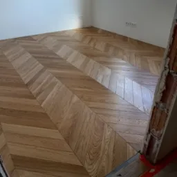 Parkiet ułożony w jodełkę francuską, z widocznymi detalami usłojenia drewna. Fragment ściany z gniazdkami elektrycznymi, okno w tle. Widoczne zabezpieczenia podłogi.