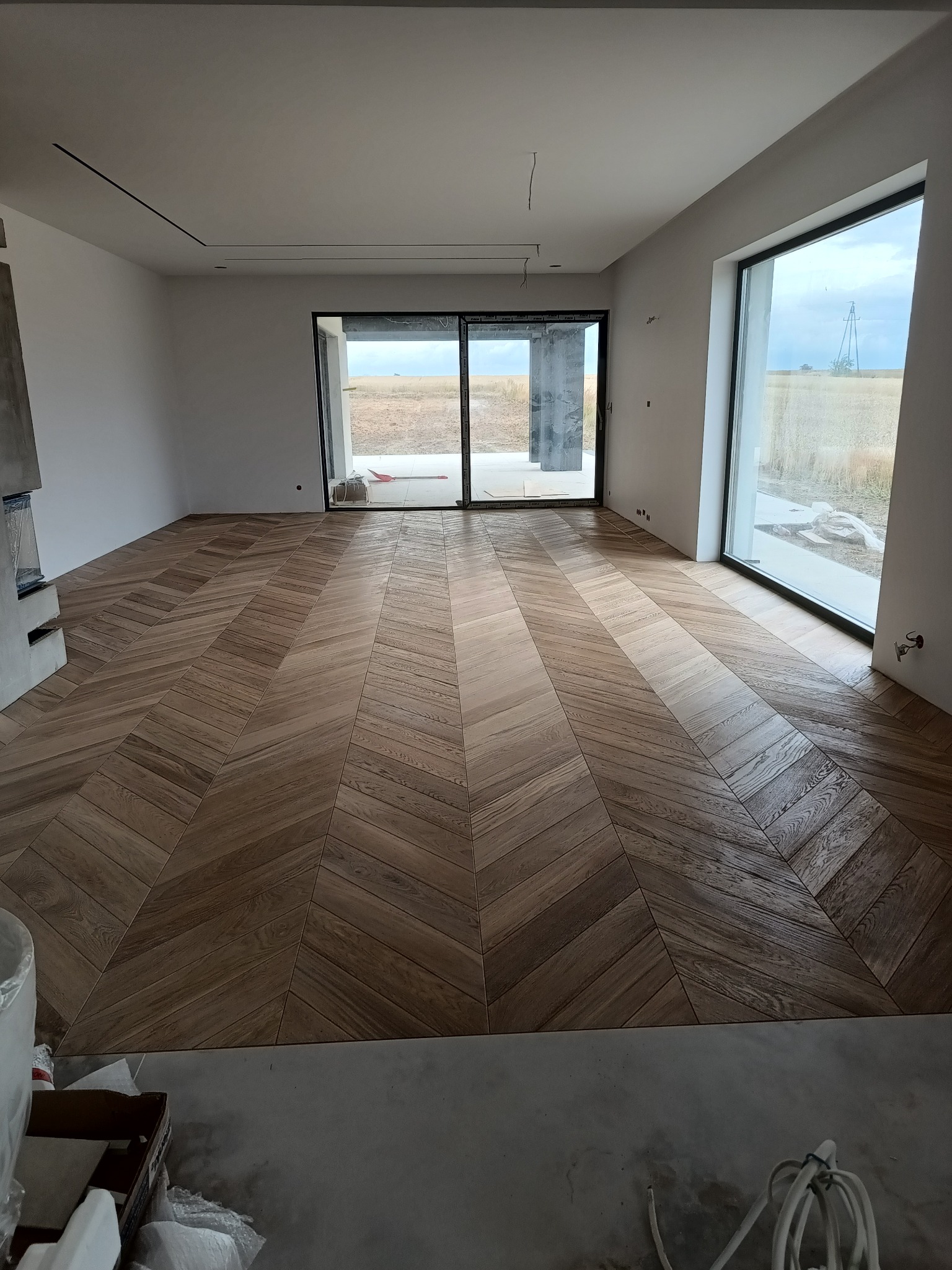 Salon z drewnianą podłogą ułożoną w jodełkę francuską, duże okna z widokiem na zewnątrz. Częściowo wykończone wnętrze.