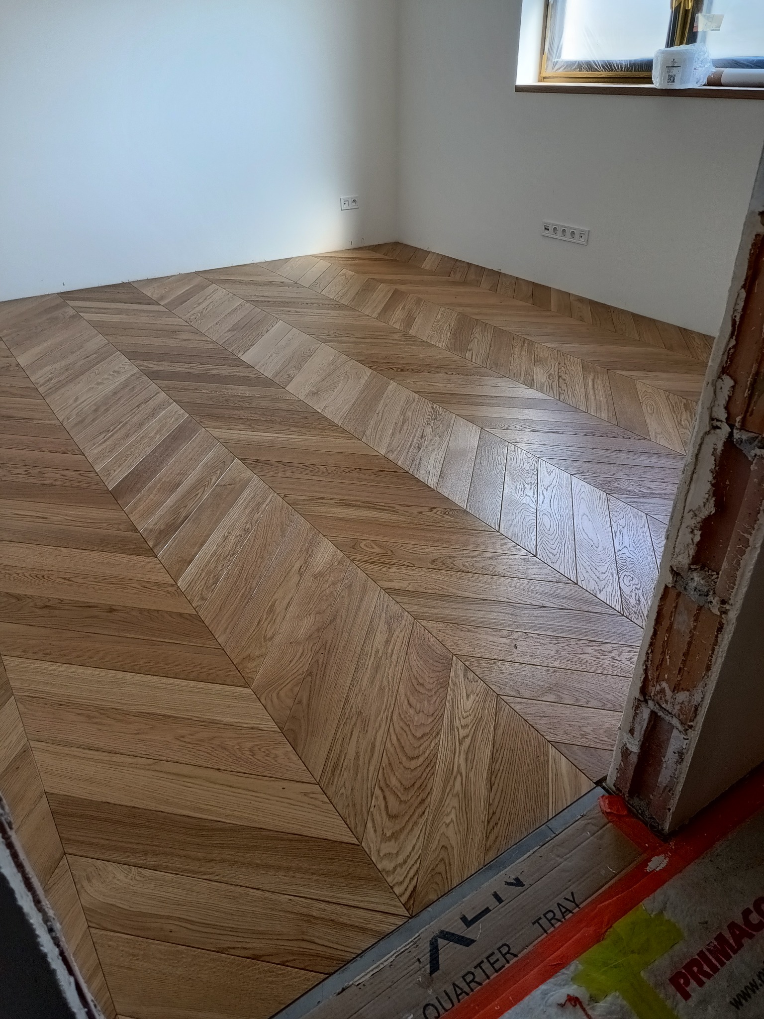 Parkiet ułożony w jodełkę francuską, z widocznymi detalami usłojenia drewna. Fragment ściany z gniazdkami elektrycznymi, okno w tle. Widoczne zabezpieczenia podłogi.