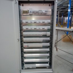 RK. - Otwarta szafa elektryczna z modułami zabezpieczeń w przemysłowym wnętrzu; widoczne kable i metalowe konstrukcje.