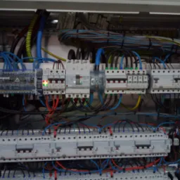 Rozdzielnia elektryczna z bezpiecznikami, przewodami i elementami sterującymi. Widoczne kable w różnych kolorach i oznaczenia na urządzeniach.