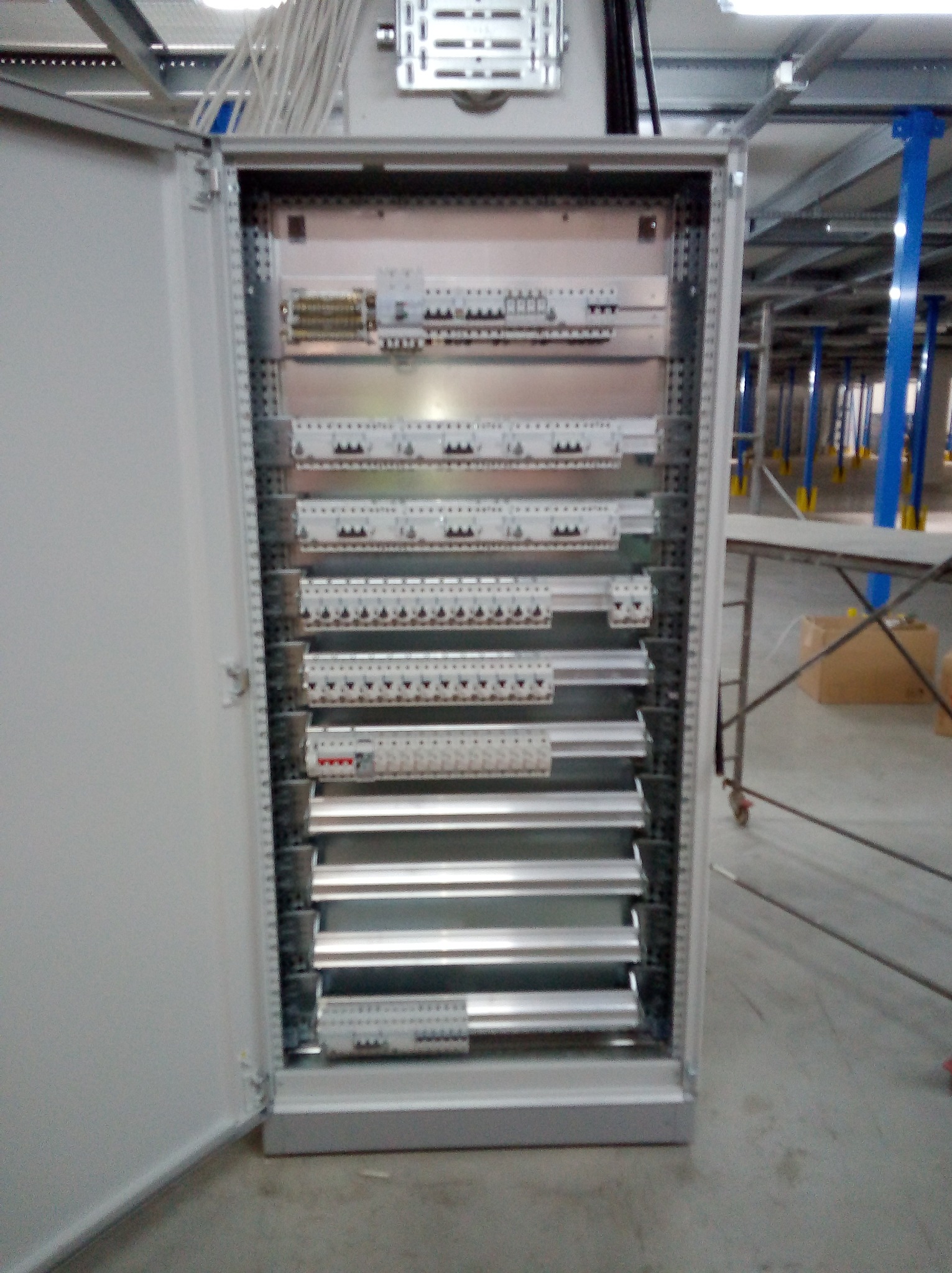 Otwarta szafa elektryczna z modułami zabezpieczeń w przemysłowym wnętrzu; widoczne kable i metalowe konstrukcje.