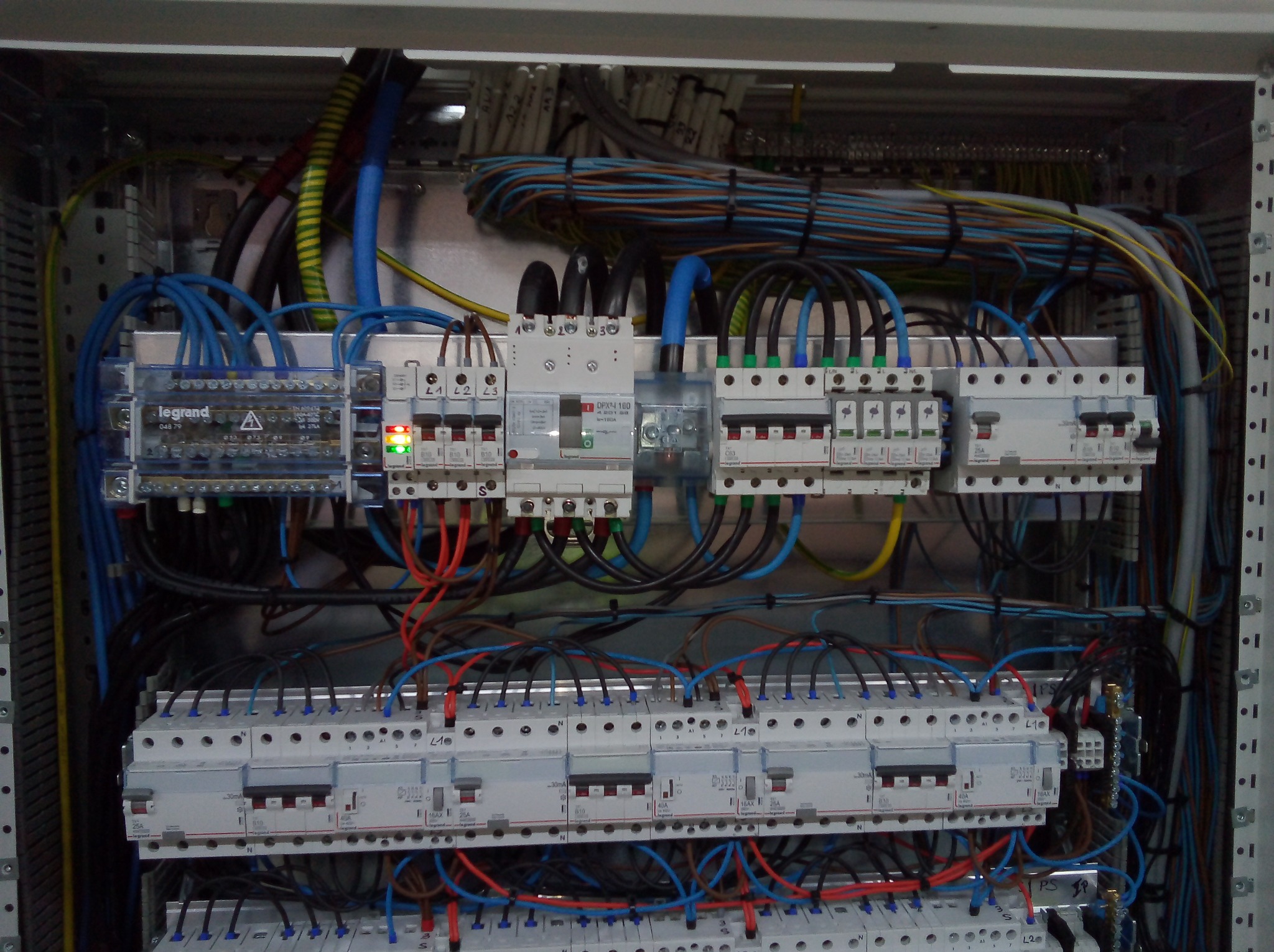 Rozdzielnia elektryczna z bezpiecznikami, przewodami i elementami sterującymi. Widoczne kable w różnych kolorach i oznaczenia na urządzeniach.