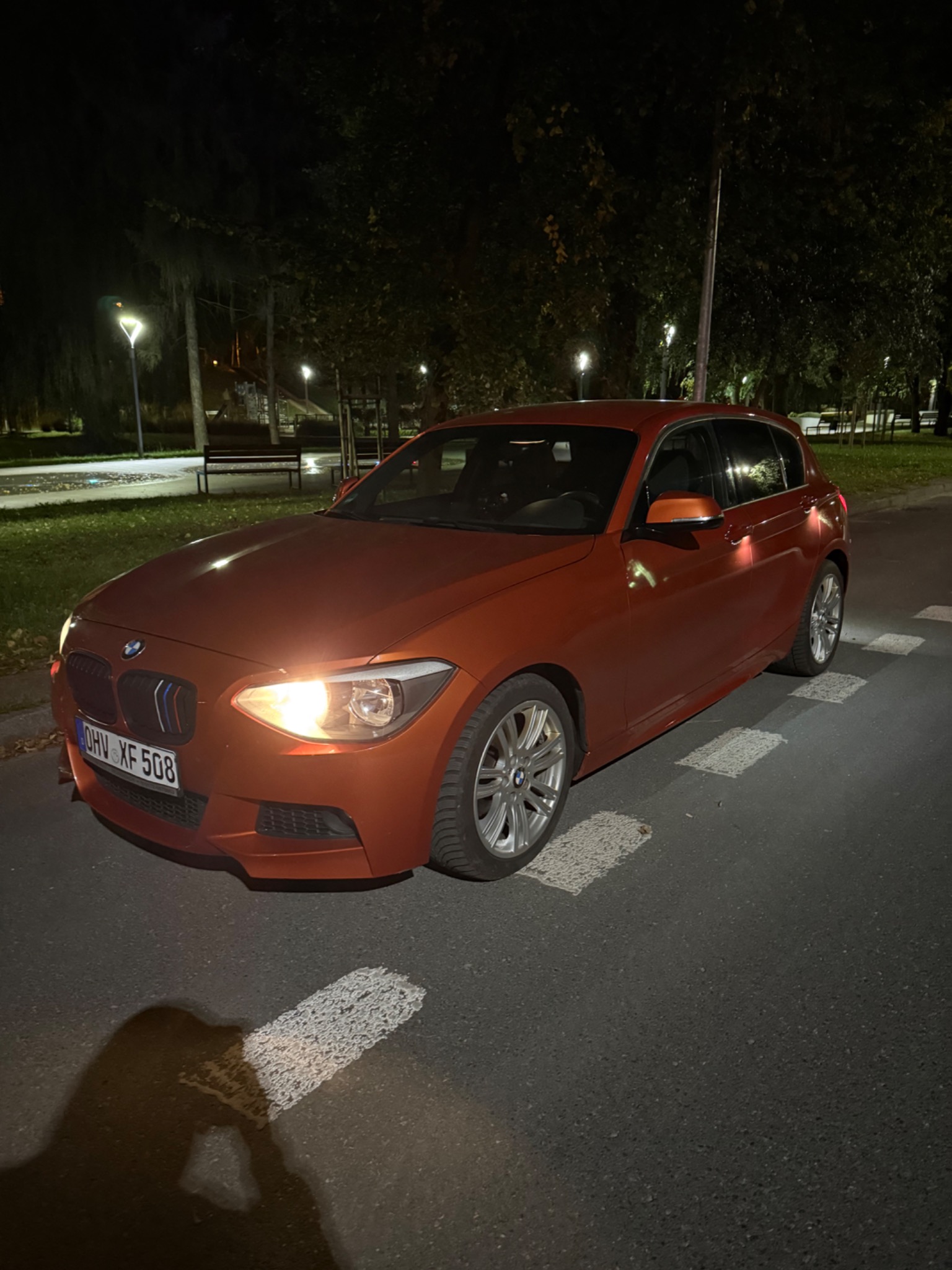 Pomarańczowy BMW serii 1 na parkingu nocą, światła włączone, widoczna tablica rejestracyjna. Auto gotowe do transportu lawetą w scenerii miejskiej.