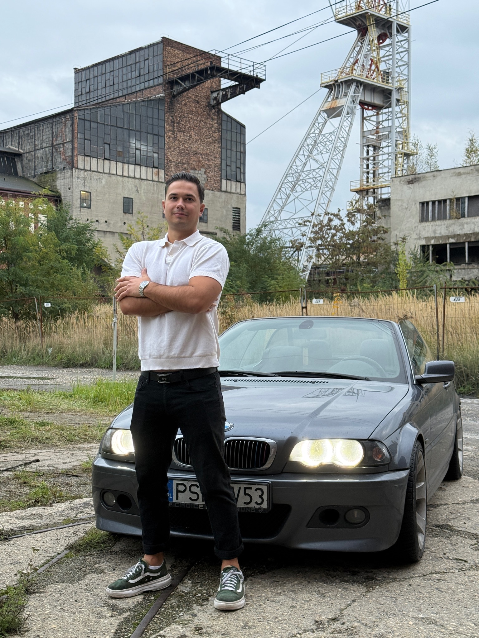 Mężczyzna w białej koszulce polo oparty o szary samochód BMW na tle opuszczonej kopalni z wieżą szybową i budynkiem maszynowni.
