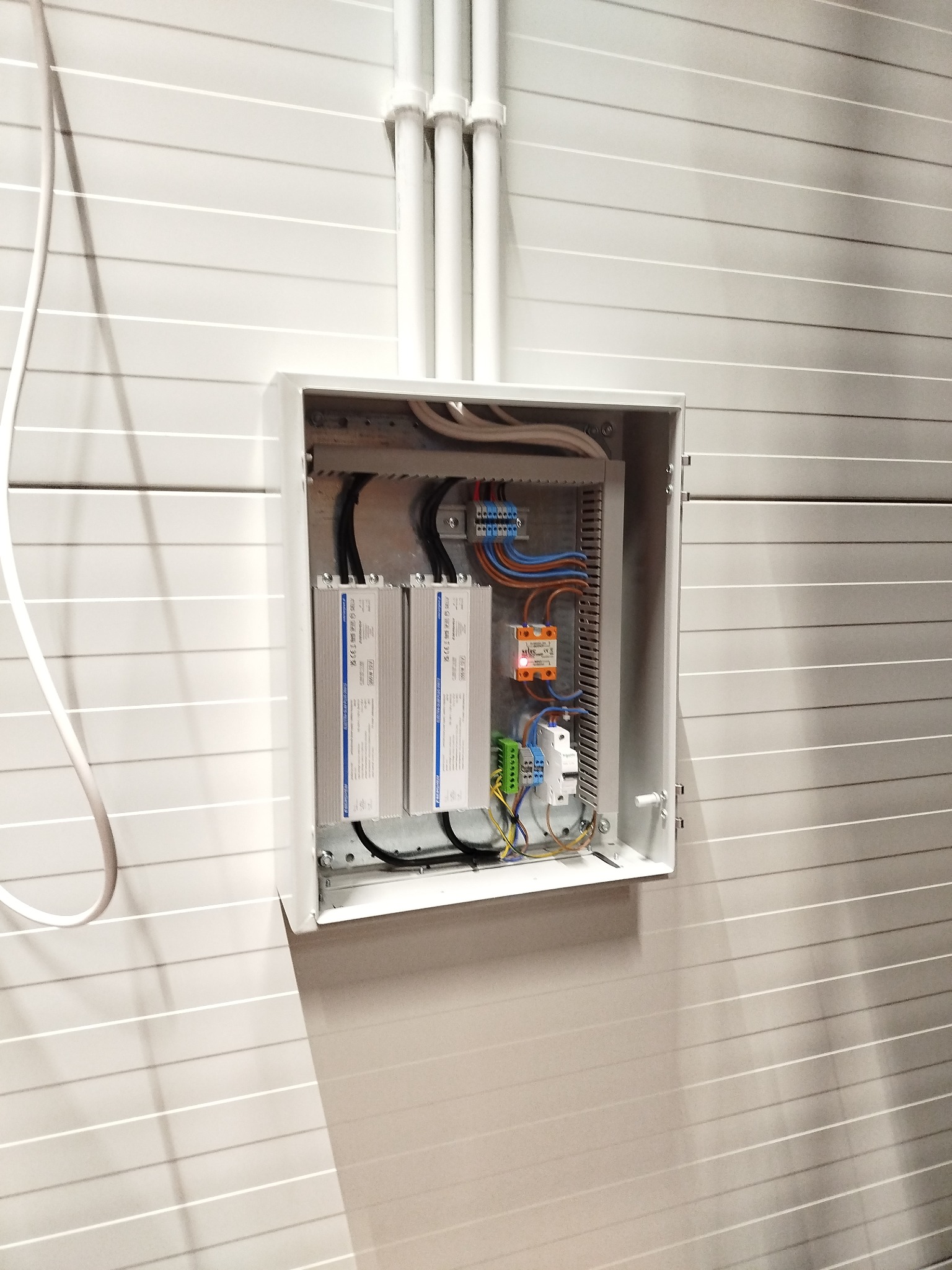 Otwarta, biała skrzynka elektryczna z przewodami, bezpiecznikami i trzema srebrnymi zasilaczami, zamontowana na ścianie z poziomymi panelami. Widoczne białe rury kablowe.