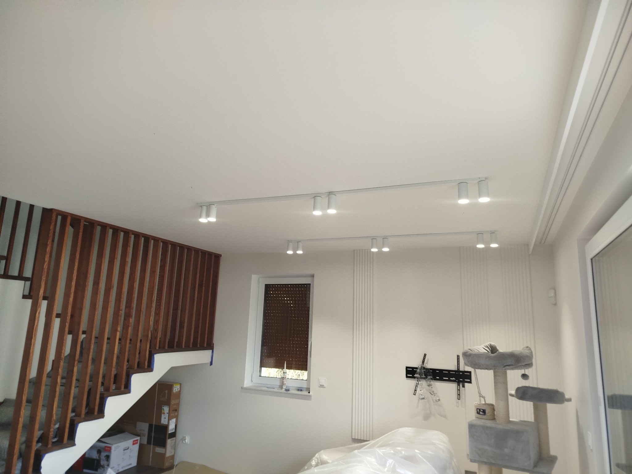 Białe reflektory szynowe zamontowane na suficie w jasnym wnętrzu z drewnianymi schodami i oknem z roletą. Widoczny uchwyt TV i drapak dla kota.