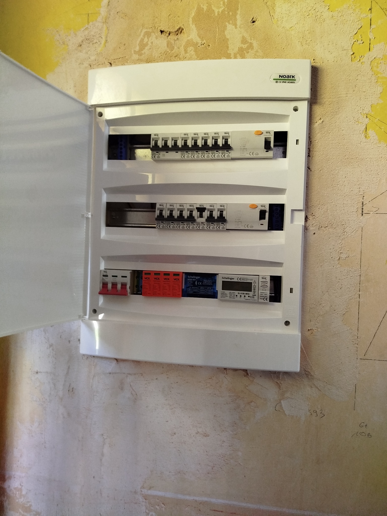 Otwarta, biała skrzynka rozdzielcza z bezpiecznikami i licznikiem na ścianie z widocznymi śladami po malowaniu. Widoczne bezpieczniki, liczniki i oznaczenia producentów.
