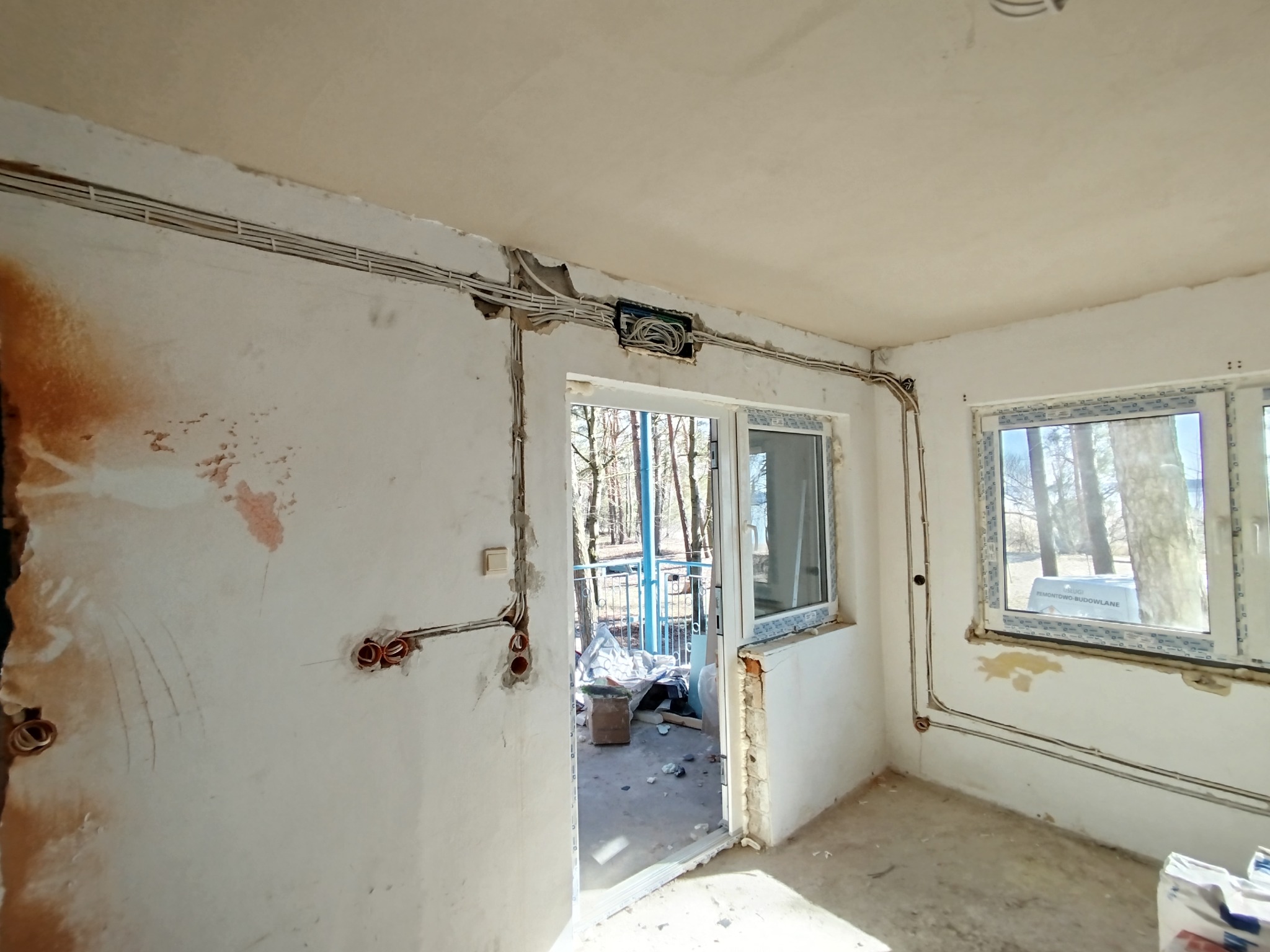Wnętrze w trakcie remontu z widoczną instalacją elektryczną: kable prowadzone po ścianach, puszka elektryczna nad oknem balkonowym. Surowy stan wykończenia.