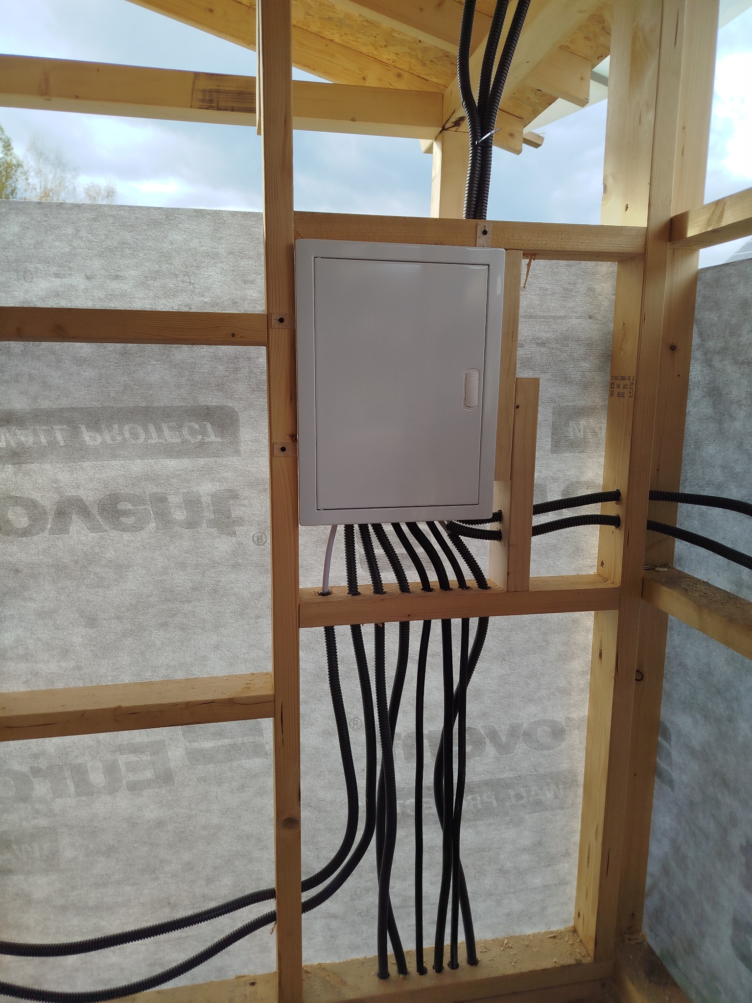 Biała skrzynka elektryczna z podłączonymi czarnymi kablami w korytkach, zamontowana w drewnianej konstrukcji szkieletowej budynku w trakcie budowy.