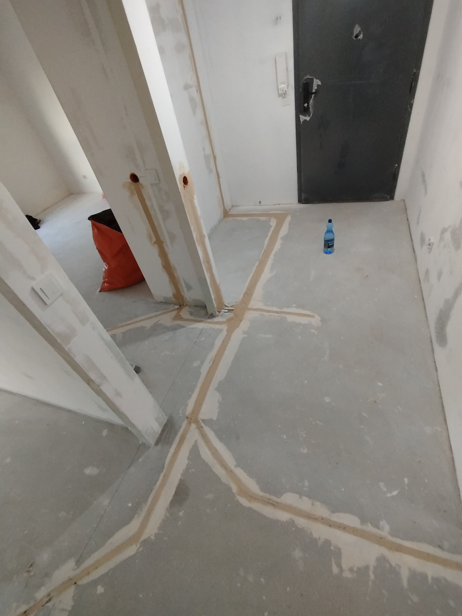 Przygotowanie podłoża pod maty grzewcze: widoczne ślady po kleju na podłodze w surowym wnętrzu, wyznaczające obszar montażu. W tle torba budowlana i butelka wody.