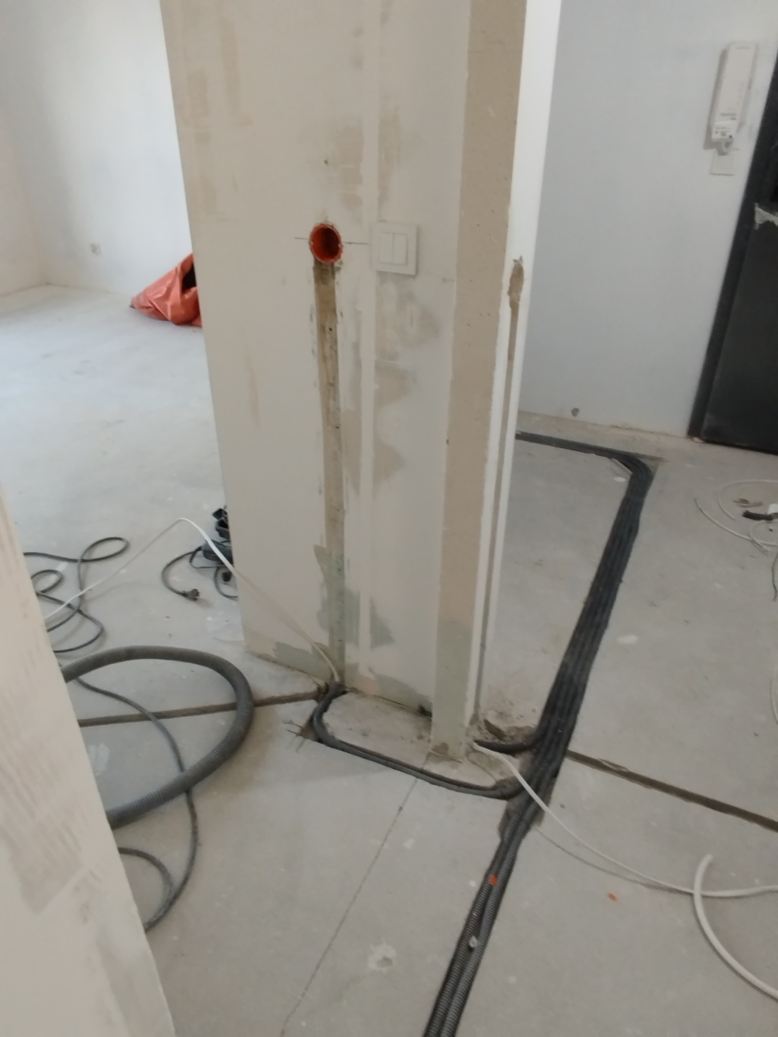 Wnętrze w trakcie prac elektrycznych: kable w peszlach ułożone w bruzdach podłogi i ściany, puszka instalacyjna w ścianie, widoczne przewody. Etap prac przed zalaniem posadzki.