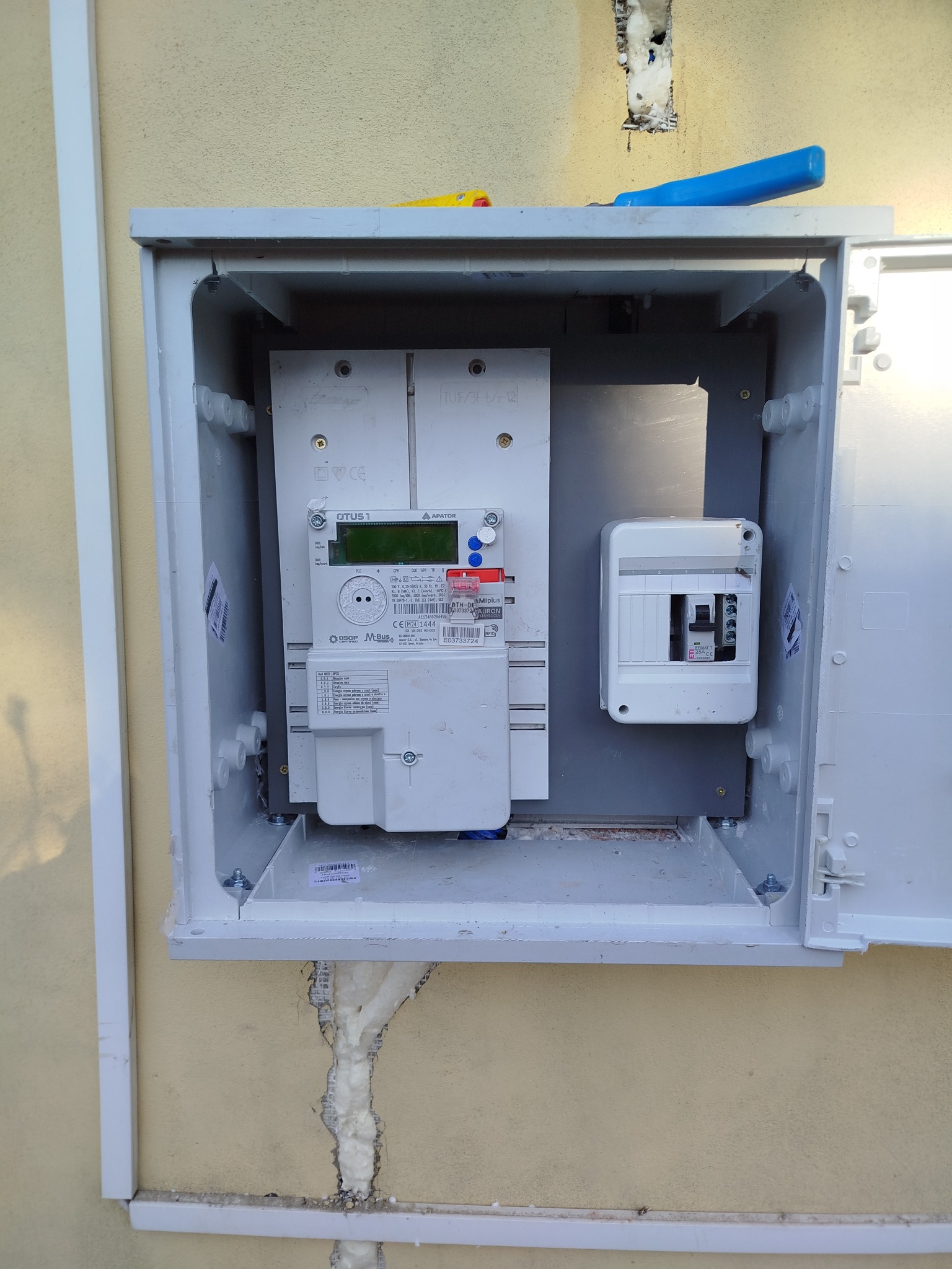 Otwarta skrzynka z licznikiem elektrycznym i bezpiecznikami na elewacji budynku, widoczne narzędzia na górze. Instalacja zabezpieczona pianką montażową.