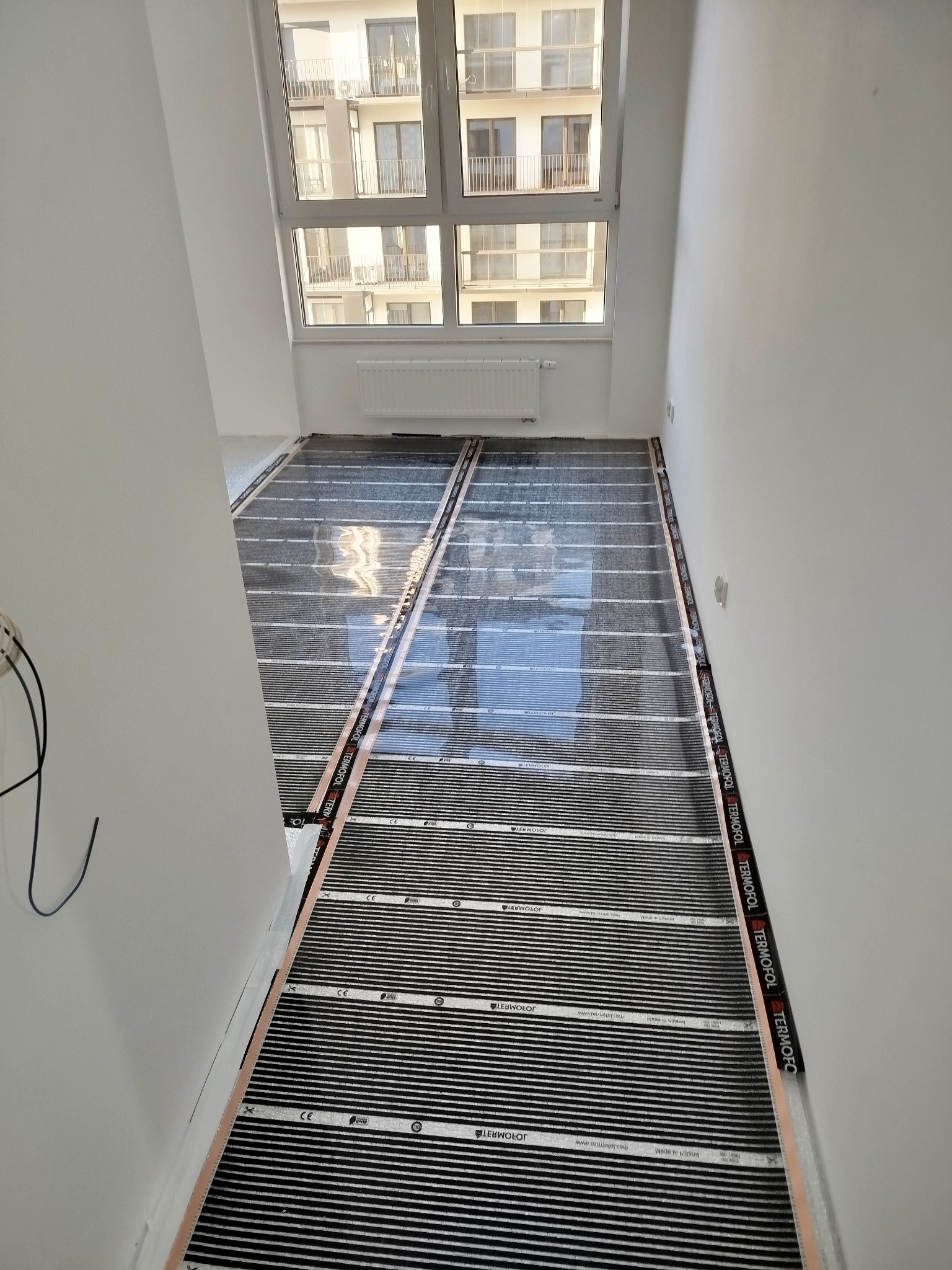 Rozłożona mata grzewcza Termofol na podłodze w jasnym wnętrzu, przygotowana do montażu. Widoczne okno z widokiem na budynek mieszkalny.