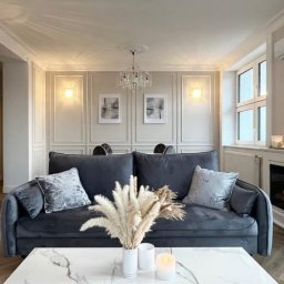 Metr za metrem - Elegancki salon w odcieniach szarości z sofą, marmurowym stolikiem, dekoracyjnymi panelami ściennymi i kominkiem elektrycznym. Całość dopełnia stylowy żyrandol.
