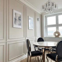 Metr za metrem - Elegancka jadalnia w stylu glamour: panele ścienne, parkiet w jodełkę, okrągły stół z chromowanymi nogami i kryształowy żyrandol. Jasne wnętrze z dekoracjami.