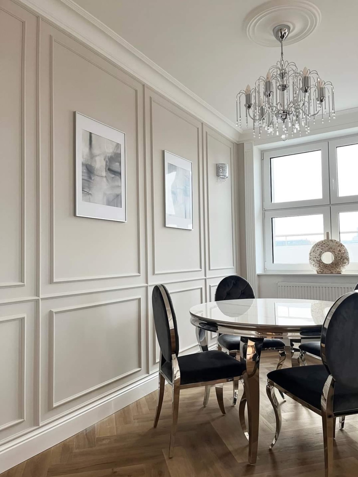 Elegancka jadalnia w stylu glamour: panele ścienne, parkiet w jodełkę, okrągły stół z chromowanymi nogami i kryształowy żyrandol. Jasne wnętrze z dekoracjami.