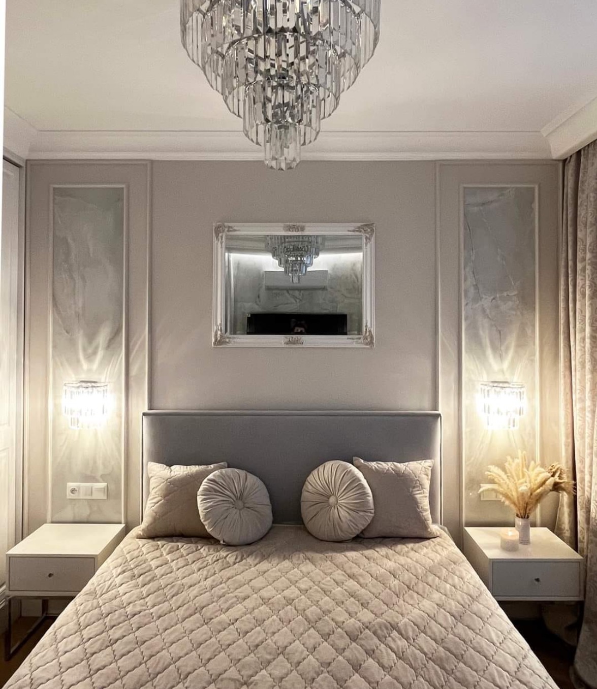 Elegancka sypialnia w stonowanej kolorystyce: pikowana narzuta, okrągłe poduszki, kryształowy żyrandol i lustro nad łóżkiem. Dwa kinkiety po bokach.