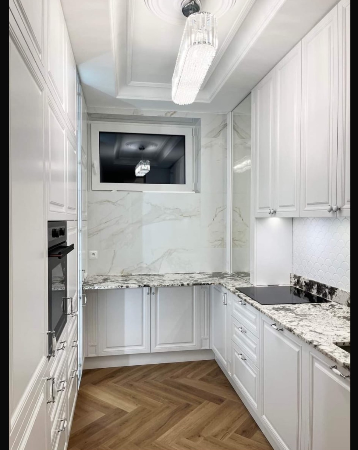 Elegancka, biała kuchnia z drewnianą podłogą w jodełkę i marmurowym blatem. Okno odbija kryształowy żyrandol, a oświetlenie punktowe podkreśla detale.