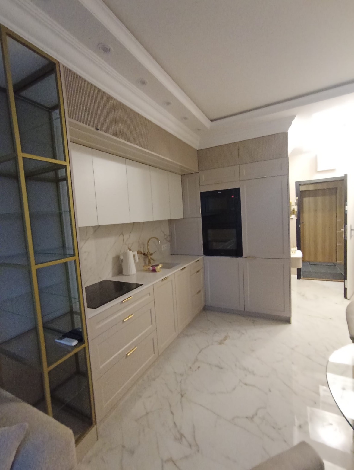 Elegancka kuchnia z beżowymi szafkami i złotymi detalami. Marmurowa podłoga i ściana dodają luksusowego charakteru. Widoczny przeszklony regał.