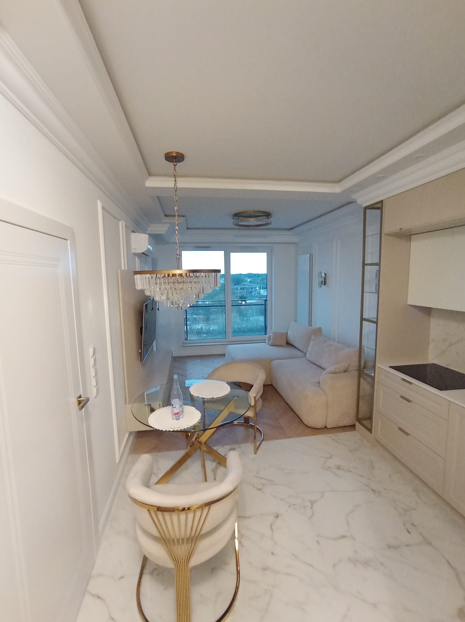 Jasne, eleganckie wnętrze: marmurowa podłoga, okrągły stół ze złotymi nogami, sofa w odcieniach beżu. Kryształowy żyrandol dodaje luksusu. Widok na zewnątrz przez okno.