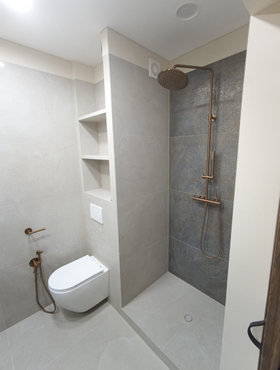 Nowoczesna łazienka z kabiną typu walk-in, szare płytki, deszczownica w kolorze miedzi, toaleta podwieszana i półki wnękowe. Minimalistyczny design.