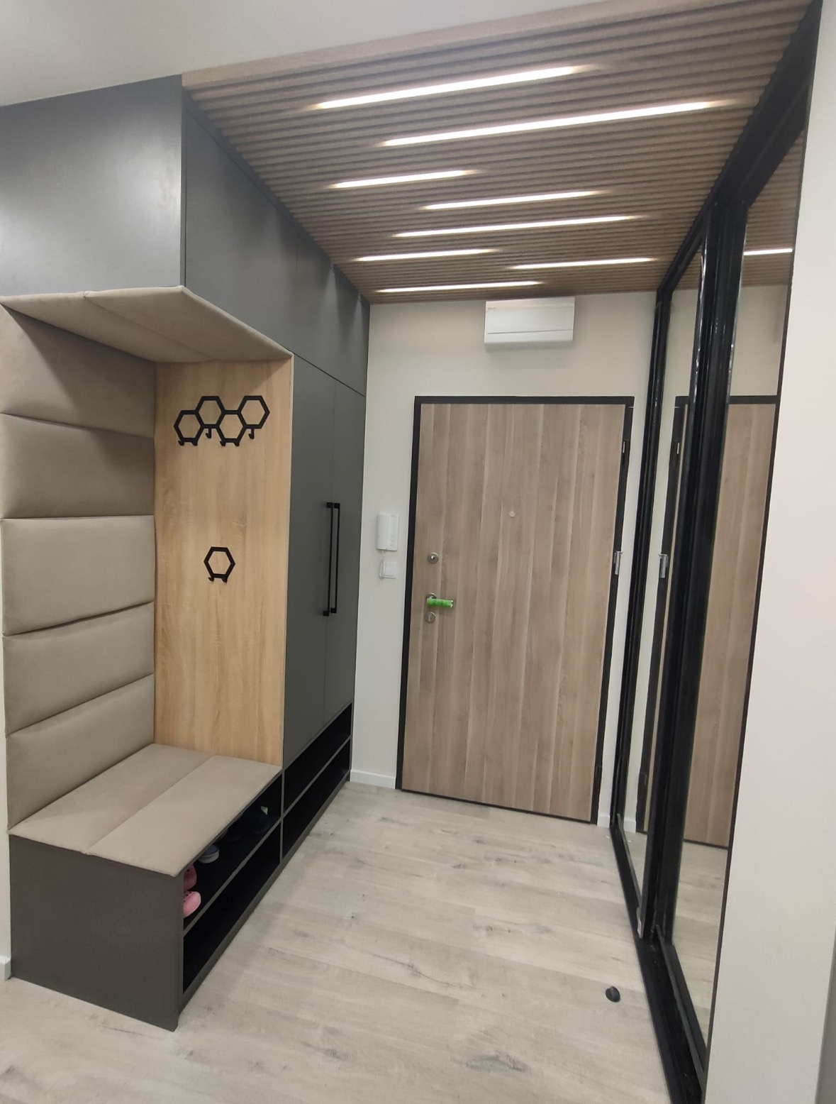 Nowoczesny przedpokój z siedziskiem, szafą w zabudowie i lustrem. Elementy drewniane z heksagonalnymi wieszakami, oświetlenie sufitowe. Minimalistyczny design.