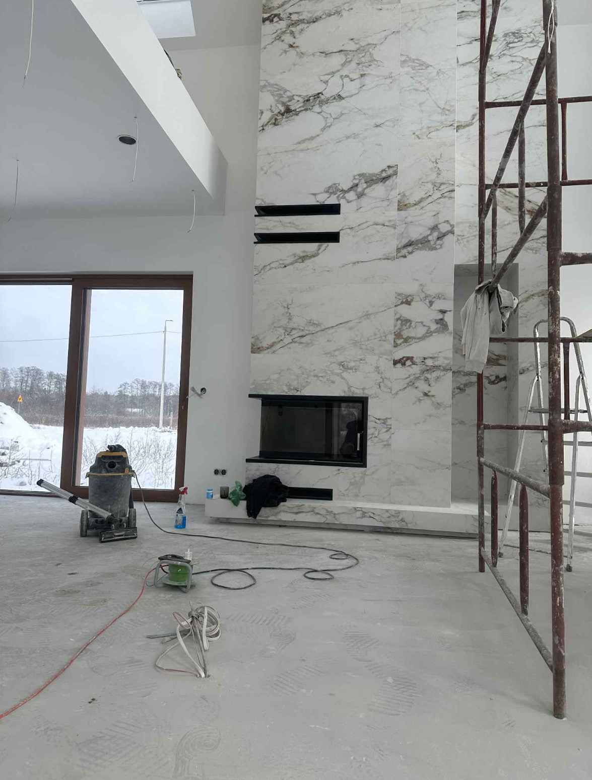 Wykończenie kominka marmurowymi płytami w nowoczesnym salonie, widoczne rusztowanie i narzędzia, wysokie okno z widokiem na zimowy krajobraz.