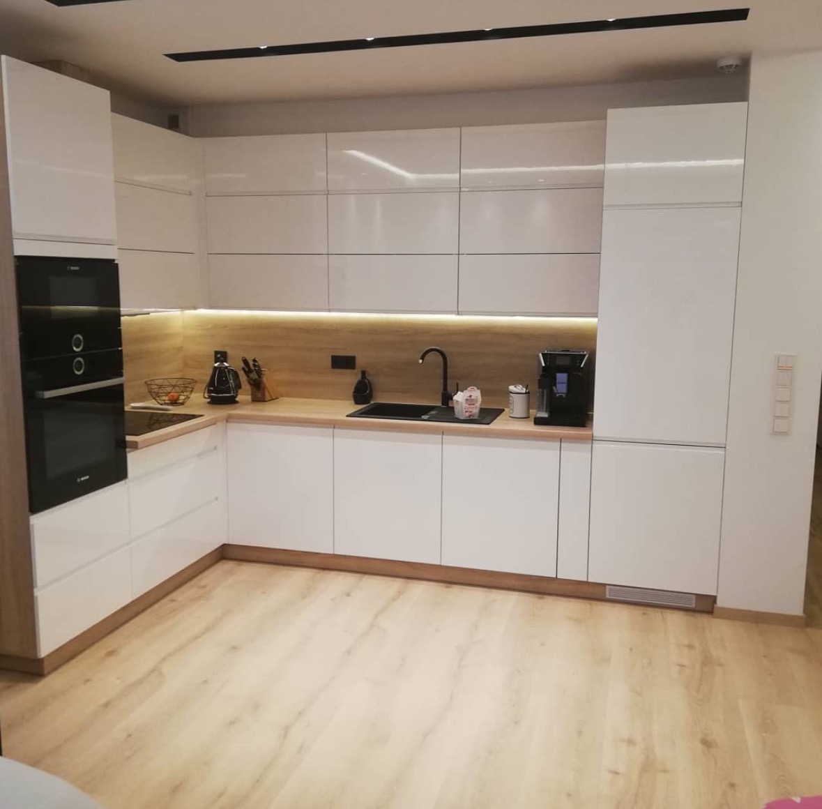 Nowoczesna kuchnia z białymi, lakierowanymi frontami i drewnianym blatem. Czarna armatura, oświetlenie LED podszafkowe. Jasna podłoga imitująca drewno.