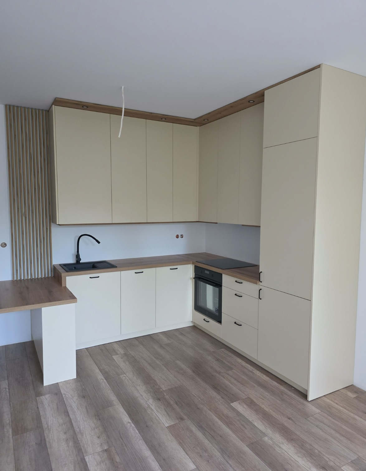 Jasna kuchnia z drewnianym blatem i czarną armaturą. Uwagę przykuwają minimalistyczne fronty szafek i drewniane panele dekoracyjne na ścianie.