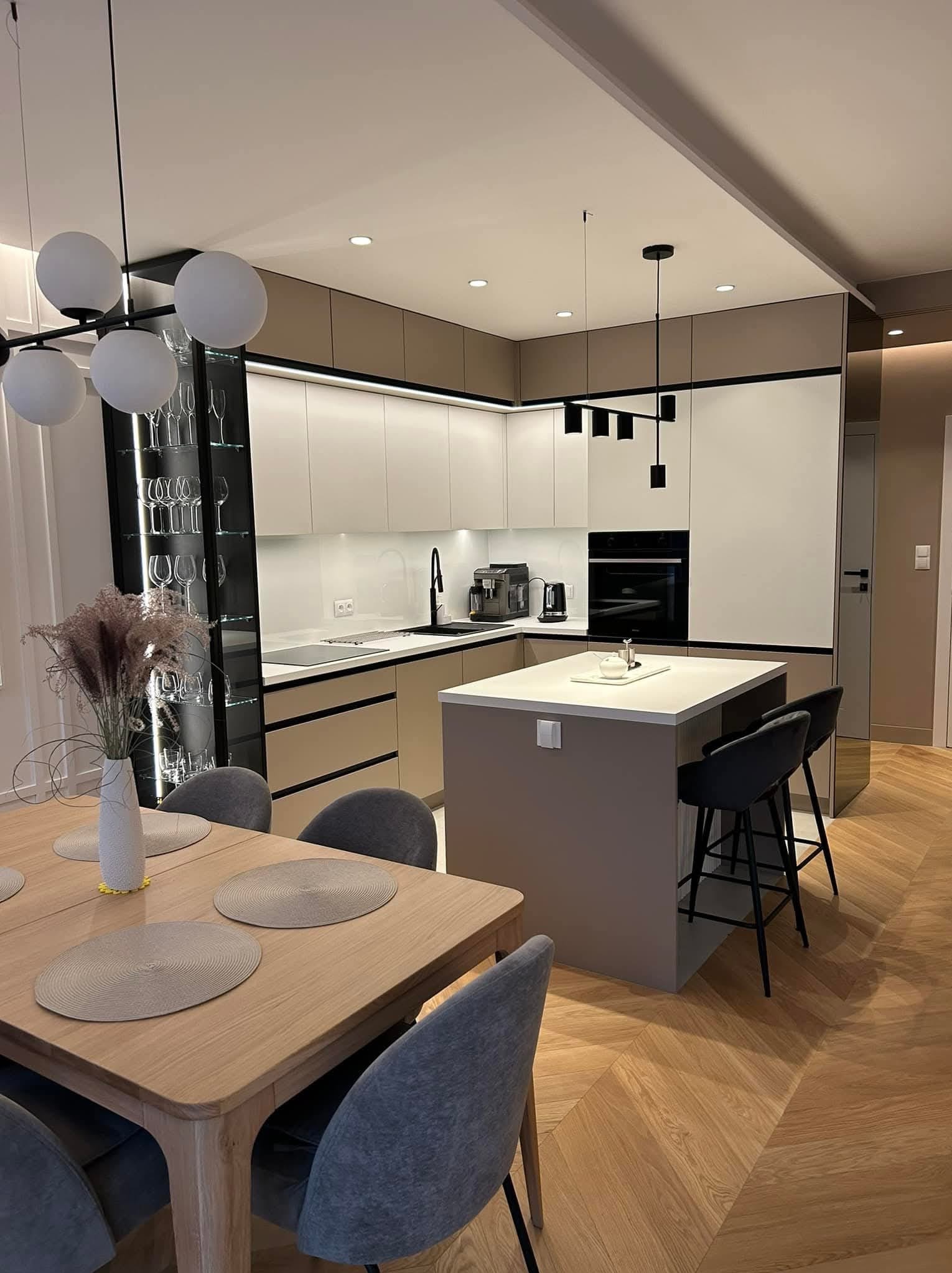 Nowoczesna kuchnia z wyspą i parkietem w jodełkę. Kremowe szafki z czarnymi akcentami, szklana witryna z kieliszkami. Stół z szarymi krzesłami i dekoracją.